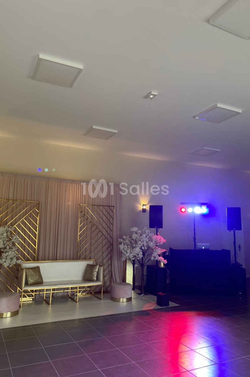 Salle de réception avec canapé, décor floral, rideaux beiges, éclairage coloré et espace DJ équipé.