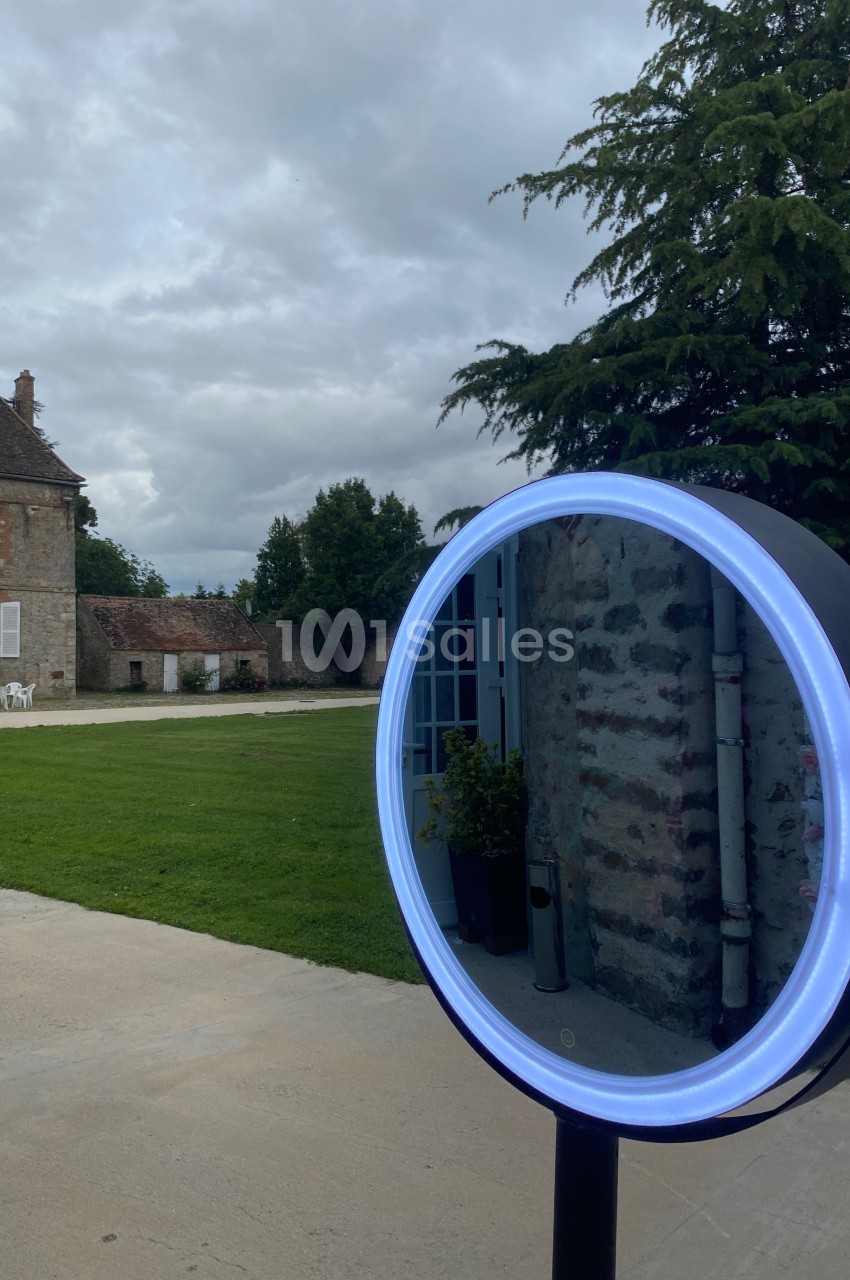 Miroir lumineux circulaire placé à l'extérieur, reflétant un bâtiment en pierre et un jardin sous un ciel nuageux.