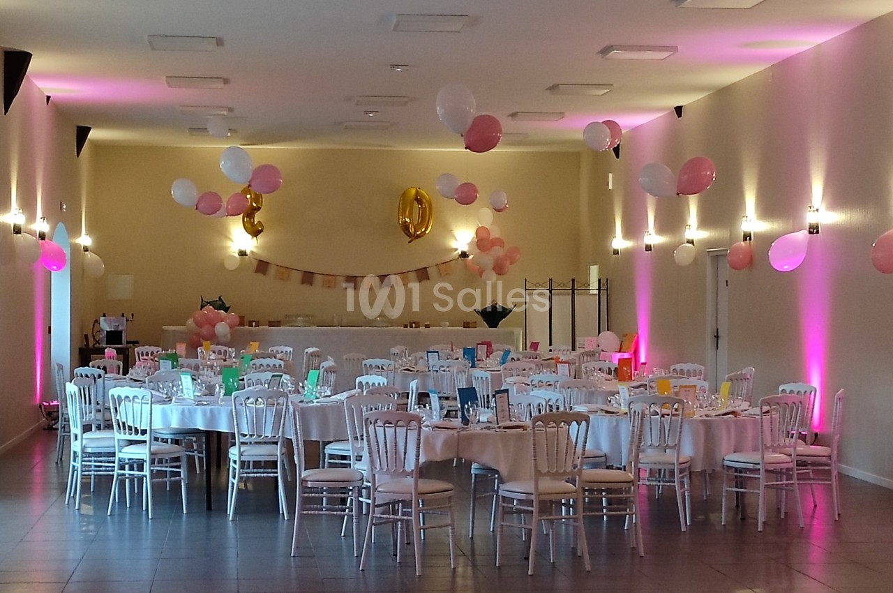Salle décorée pour une fête avec des tables rondes, nappes blanches, chaises en bois et ballons roses et blancs suspendus.