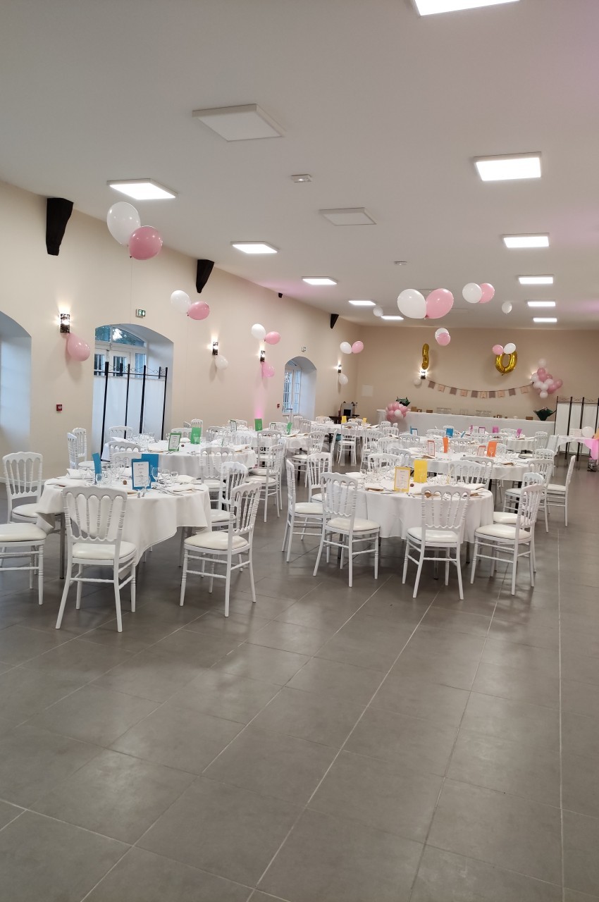 Salle décorée pour une fête, avec des tables rondes dressées, des chaises blanches et des ballons roses et blancs suspendus.