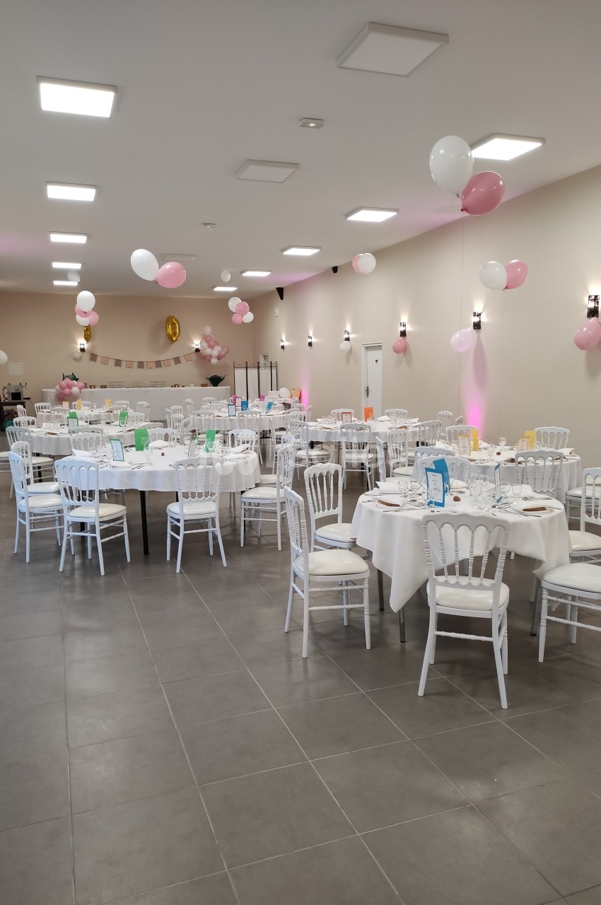 Salle décorée pour un événement avec des tables rondes dressées, des chaises blanches et des ballons roses et blancs.