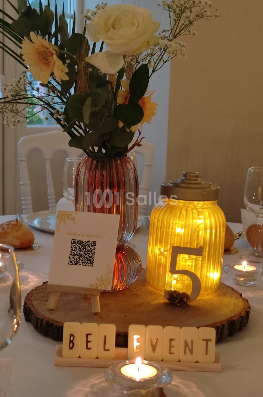 Centre de table avec fleurs, bougie LED dans un pot numéroté ’5’, QR code et lettres formant ’BEL EVENT’.