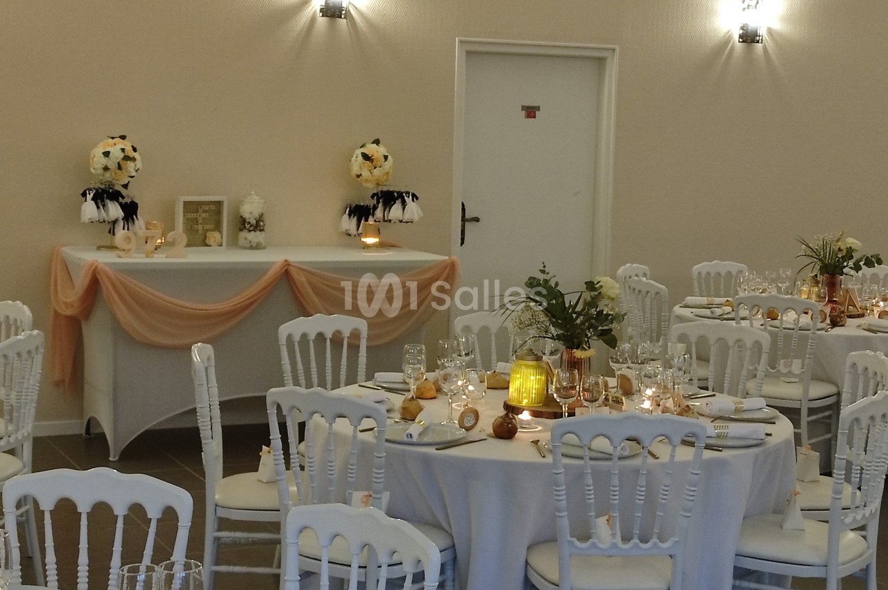 Salle de réception décorée avec des tables rondes dressées, des chaises blanches et un buffet orné de fleurs et de voilages.