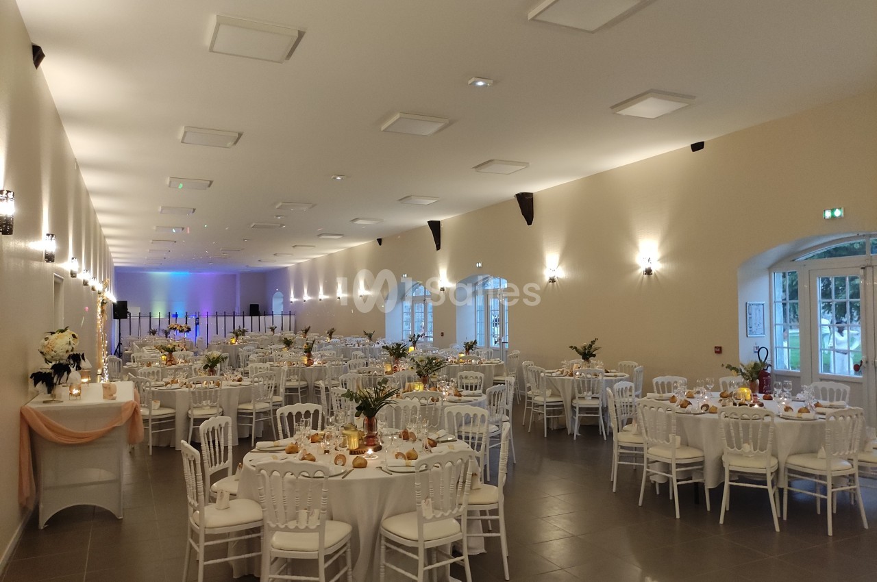 Salle de réception lumineuse avec des tables rondes décorées pour un événement, chaises blanches et éclairage chaleureux.