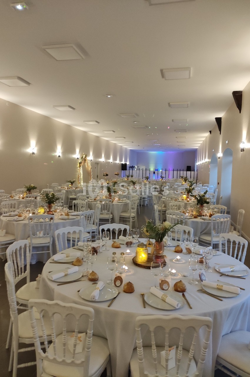 Salle de réception décorée avec des tables rondes dressées, chaises blanches et éclairage chaleureux.