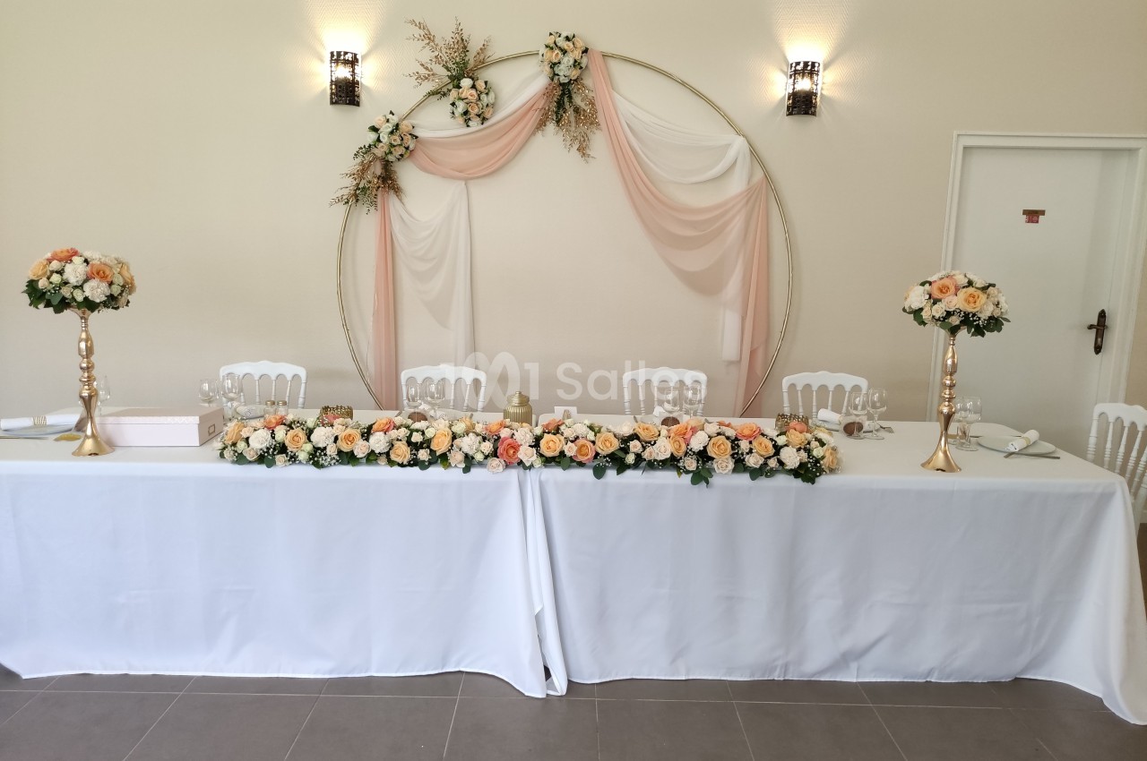 Table de réception décorée de nappes blanches, compositions florales pastel et arche ornée de voilages roses.