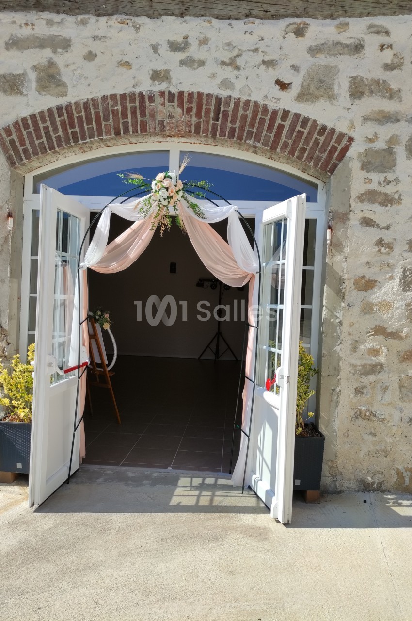 Entrée d'un bâtiment en pierre avec des portes vitrées ouvertes, décorées de voilages roses et d'une composition florale.