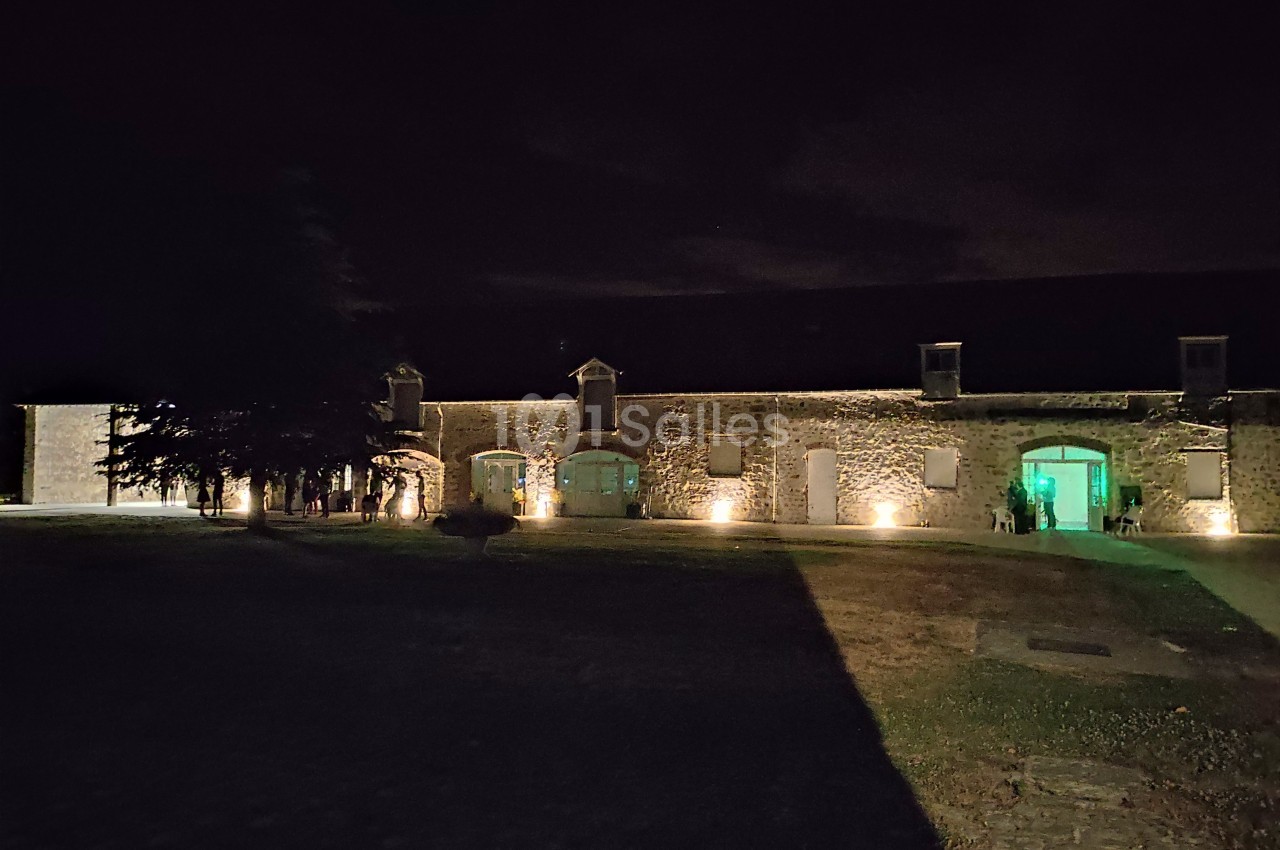 Bâtiment en pierre éclairé la nuit, avec des fenêtres illuminées et une cour partiellement ombragée.