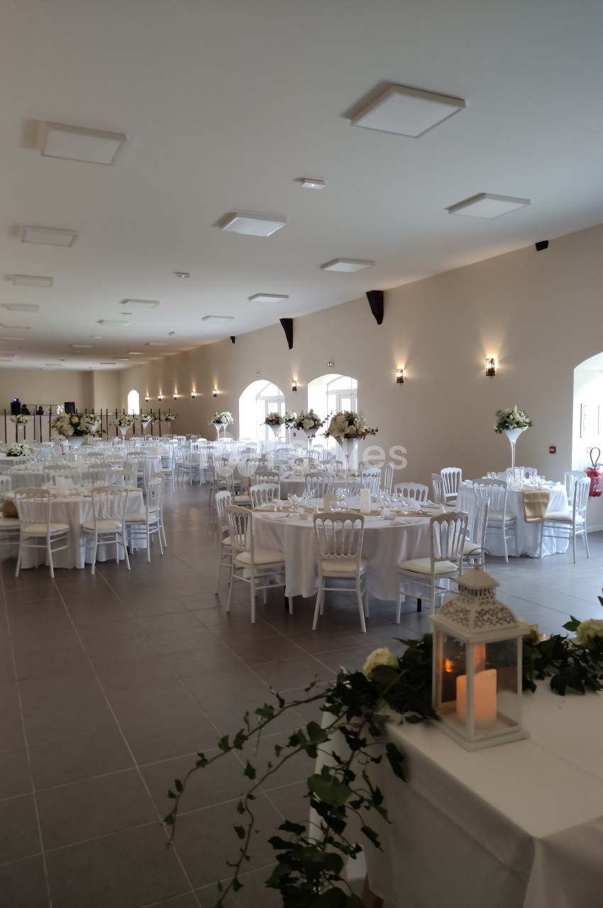 Salle de réception lumineuse avec tables rondes dressées, chaises blanches et décorations florales élégantes.