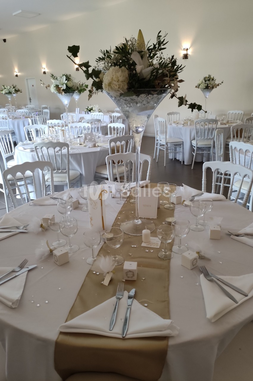 Salle de réception décorée pour un mariage, avec tables rondes dressées, nappes blanches et centres de table fleuris.
