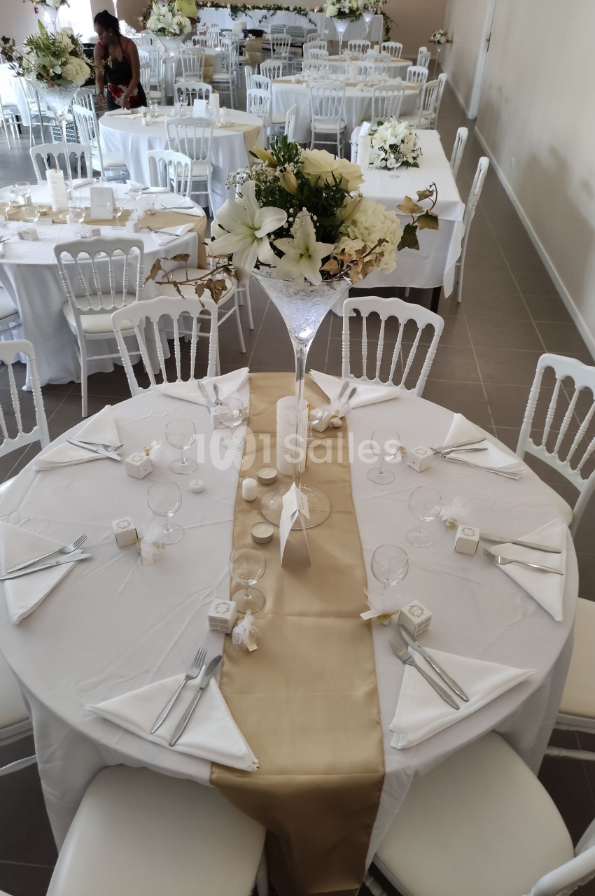 Salle de réception avec tables rondes décorées de nappes blanches, chemins de table dorés et centres de table fleuris.