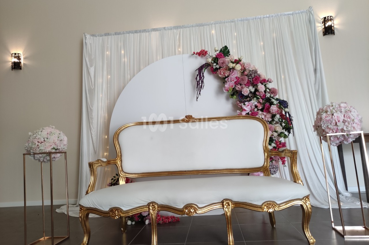 Canapé doré avec assise blanche devant un décor floral et un rideau blanc, éclairé par des appliques murales.