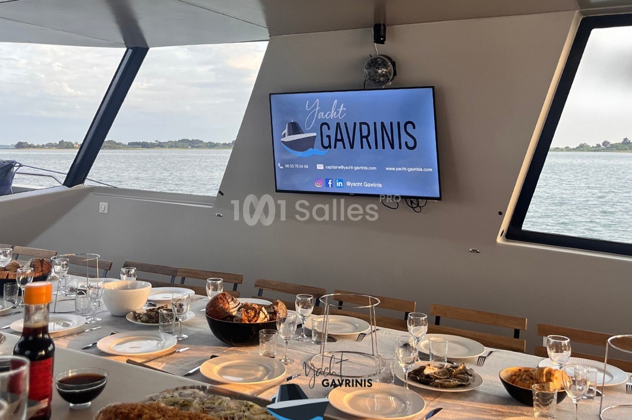 Table dressée pour un repas sur un yacht avec vue sur l'eau et un écran affichant ’Yacht Gavrinis’.
