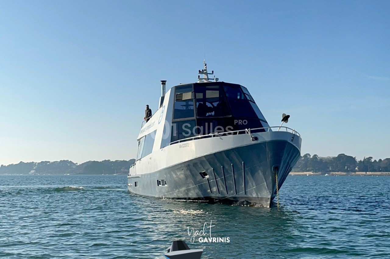 Bateau moderne naviguant sur une mer calme, avec un rivage boisé visible à l'arrière-plan.