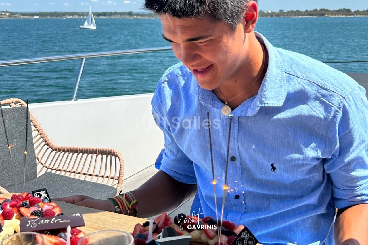 Un homme souriant assis à une table garnie de desserts, sur un bateau avec vue sur la mer et un voilier au loin.