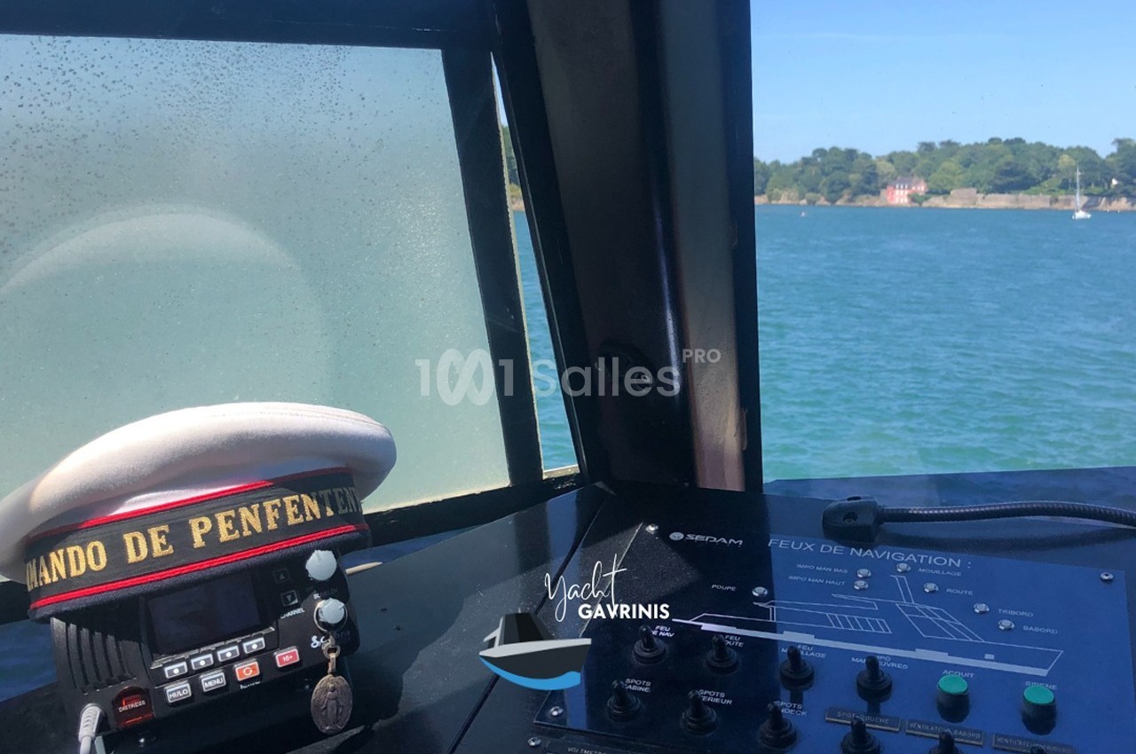 Vue depuis la cabine d'un bateau avec tableau de commande, radio et casquette posée, face à une étendue d'eau.