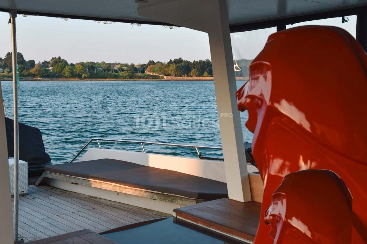 Vue depuis un bateau avec un siège orange au premier plan et un paysage côtier en arrière-plan.