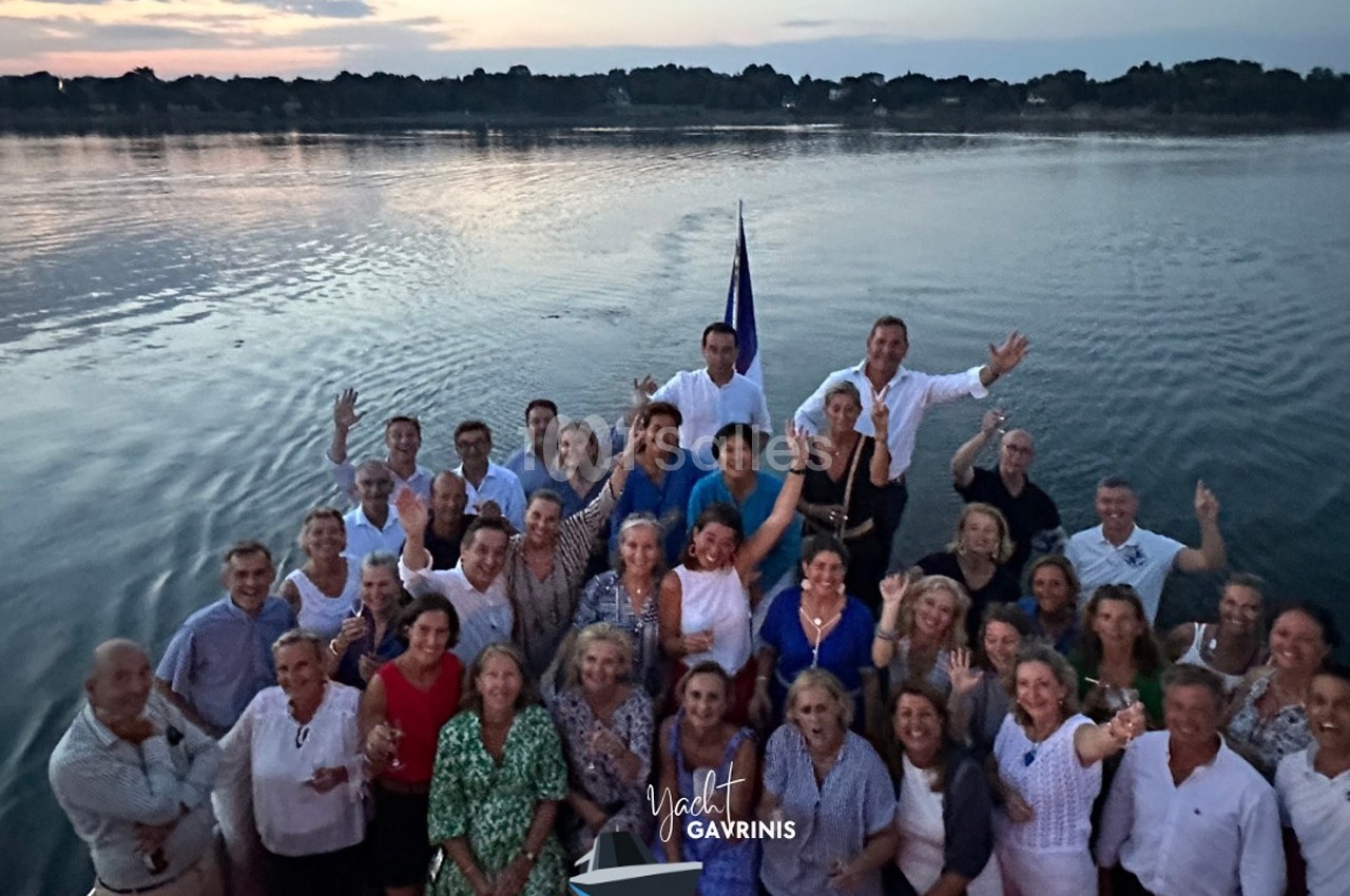 Groupe de personnes souriantes posant sur le pont d'un bateau au crépuscule, avec un paysage aquatique en arrière-plan.