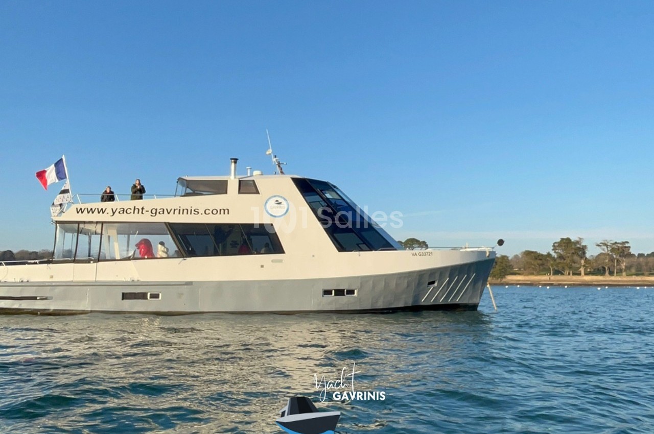 Bateau moderne naviguant sur une eau calme, avec des passagers à bord et un drapeau français à l'arrière.