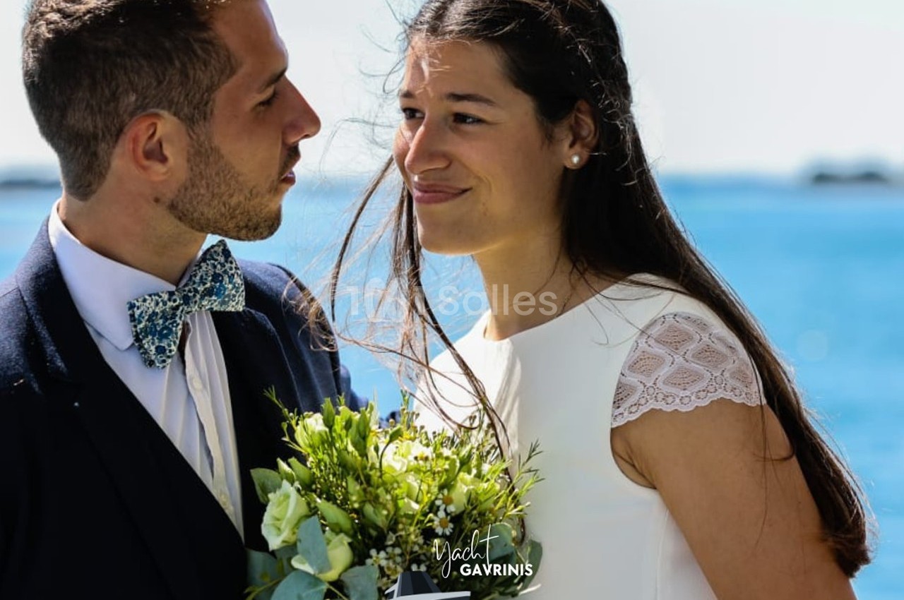 Un couple en tenue de mariage se regarde avec complicité devant un paysage marin.