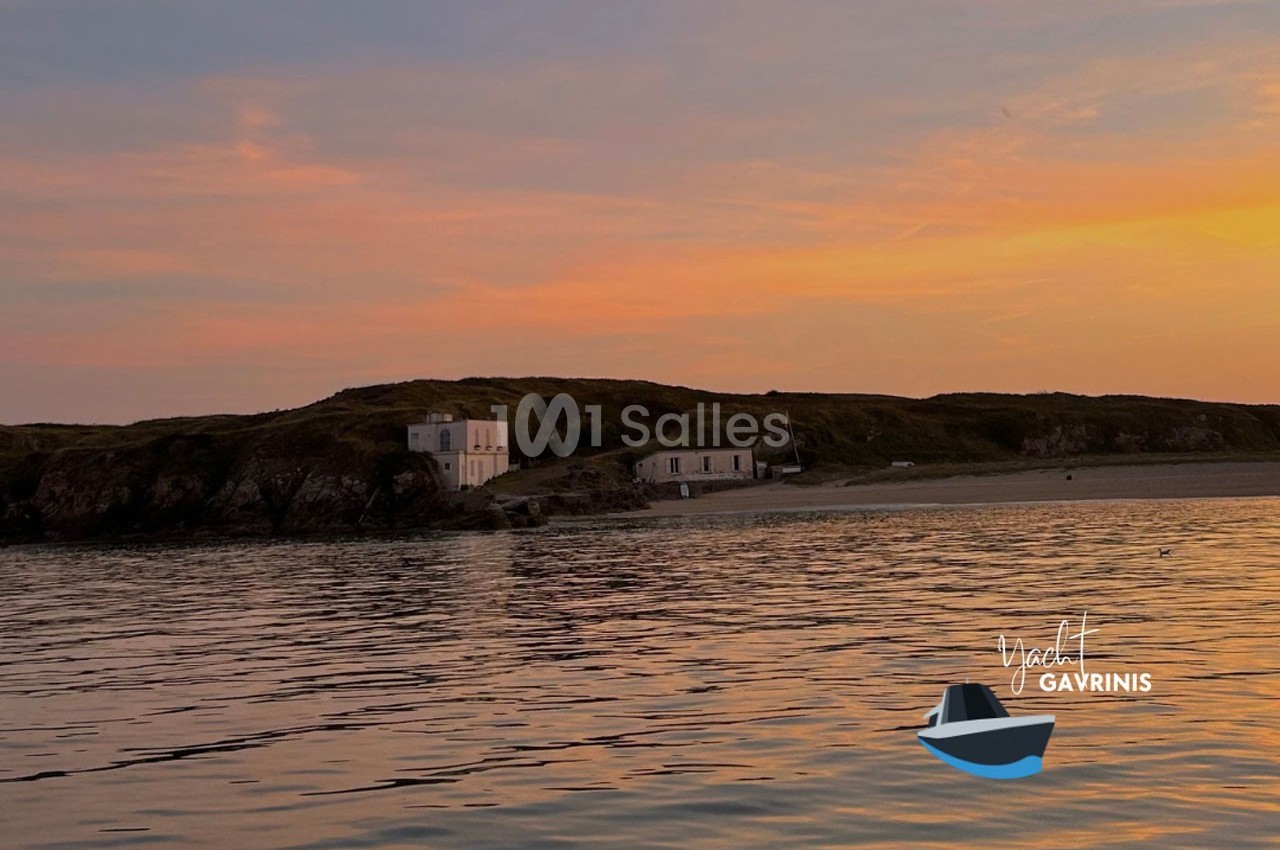Vue d'une côte rocheuse avec des bâtiments blancs au coucher du soleil, reflétée sur une mer calme.
