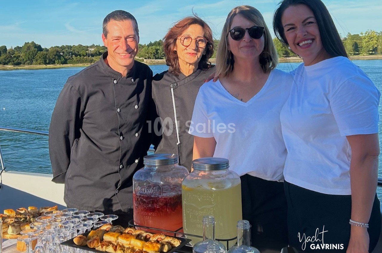 Quatre personnes souriantes devant une table dressée avec boissons et amuse-bouches, sur un bateau près d'un plan d'eau.