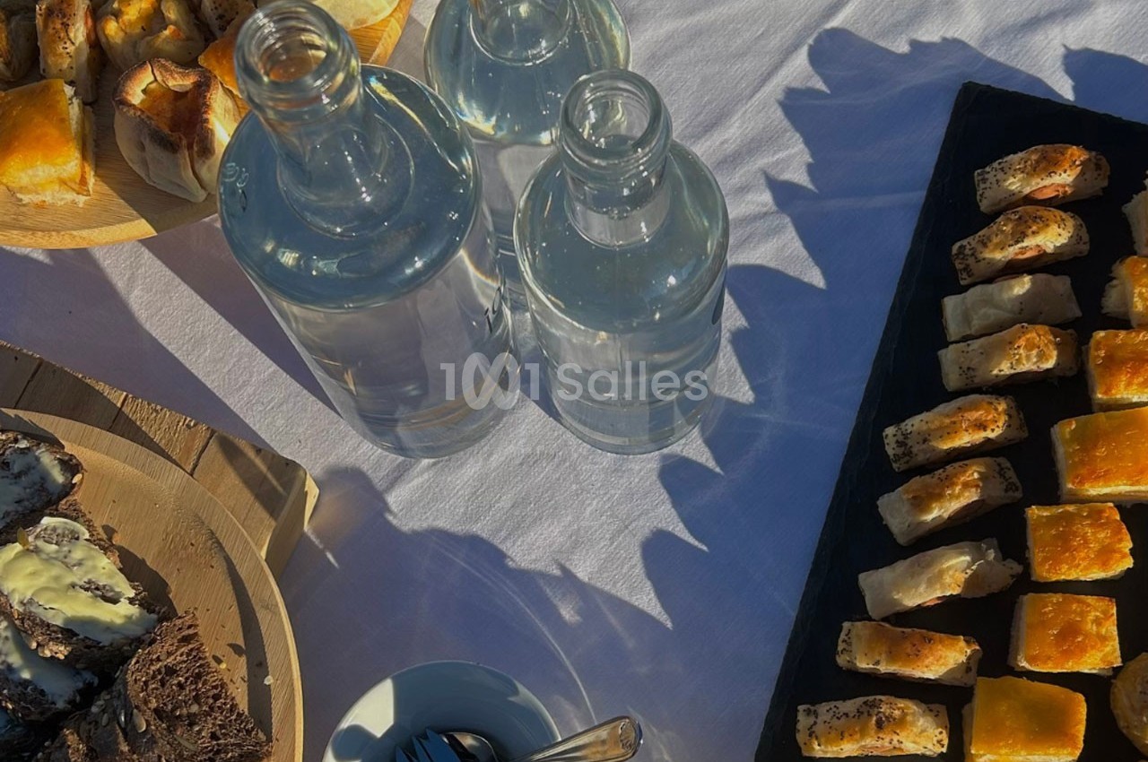 Plateaux de bouchées salées, bouteilles d'eau et couverts sur une nappe blanche en extérieur, éclairés par le soleil.