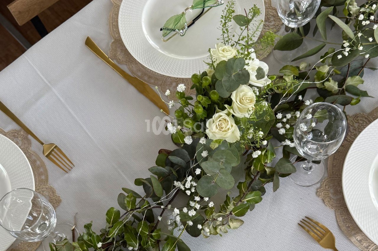 Table dressée avec une nappe blanche, couverts dorés, assiettes blanches et un centre de table floral avec des roses…