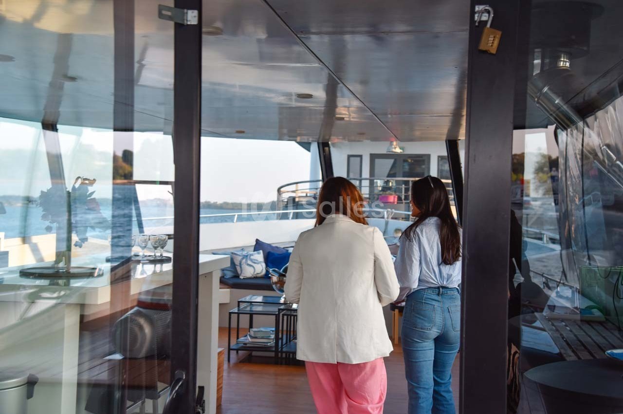 Deux femmes discutent à l'intérieur d'un bateau, avec vue sur un espace aménagé et l'eau à l'extérieur.
