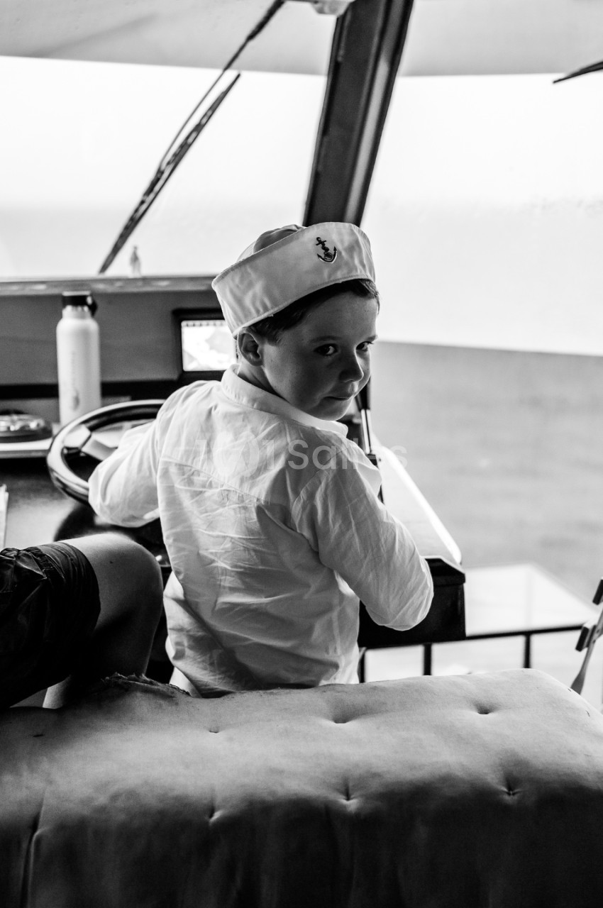 Un enfant portant une casquette de marin est assis à la barre d'un bateau, regardant par-dessus son épaule.