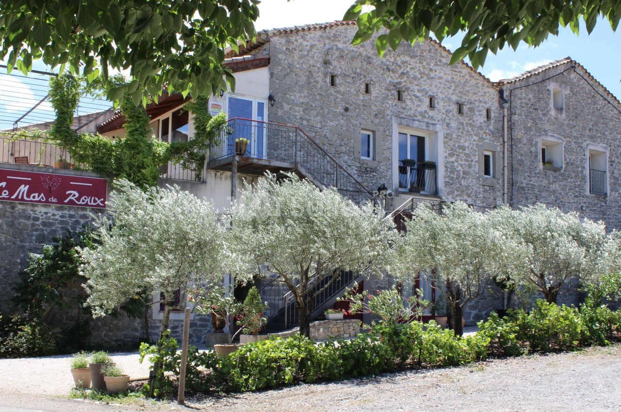 Façade d'une bâtisse en pierre entourée d'arbres et de végétation, avec un panneau indiquant ’Le Mas de Roux’.