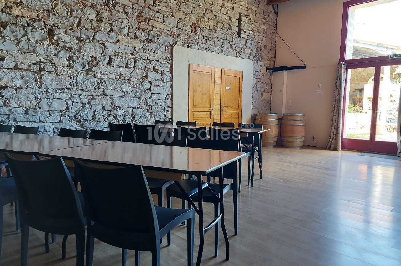 Salle lumineuse avec murs en pierre, tables et chaises noires, barriques en bois et grande porte vitrée.
