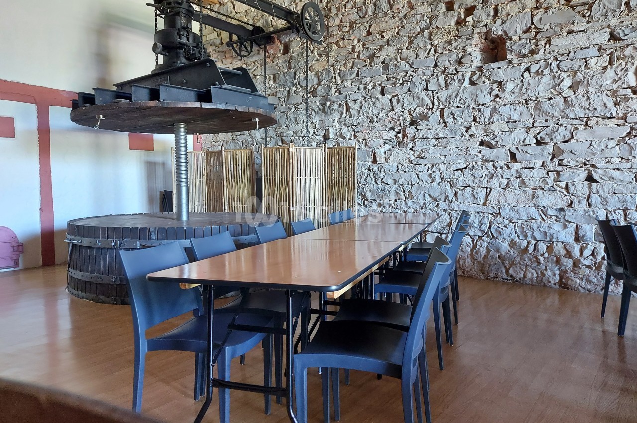 Salle avec une longue table en bois entourée de chaises noires, décorée d'un ancien pressoir et de murs en pierre.