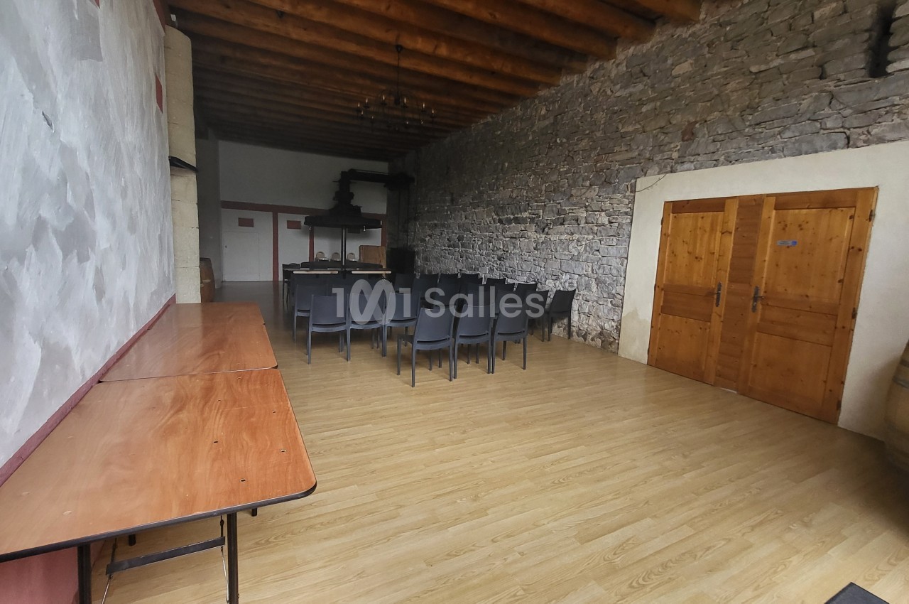 Salle avec murs en pierre et plafond en bois, équipée de chaises noires, tables en bois et tonneaux dans un coin.