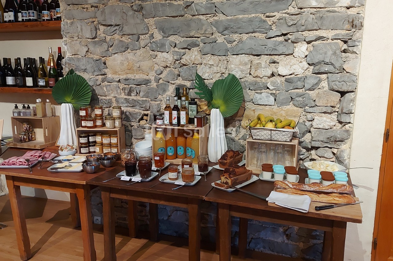 Buffet avec confitures, jus, viennoiseries, charcuteries et fruits, disposé sur des tables en bois devant un mur en pierre.