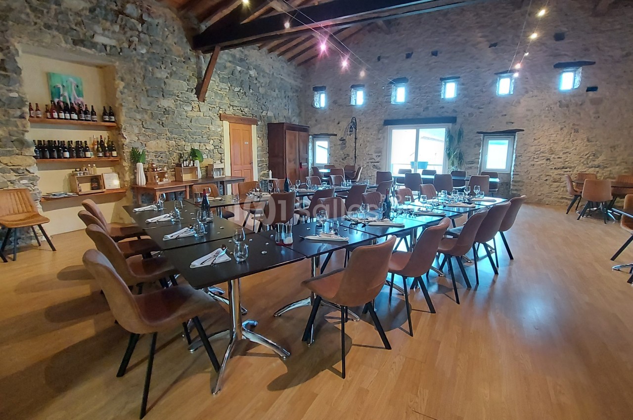 Salle en pierre avec tables dressées, chaises en cuir, étagères de bouteilles et lumière naturelle provenant des fenêtres.