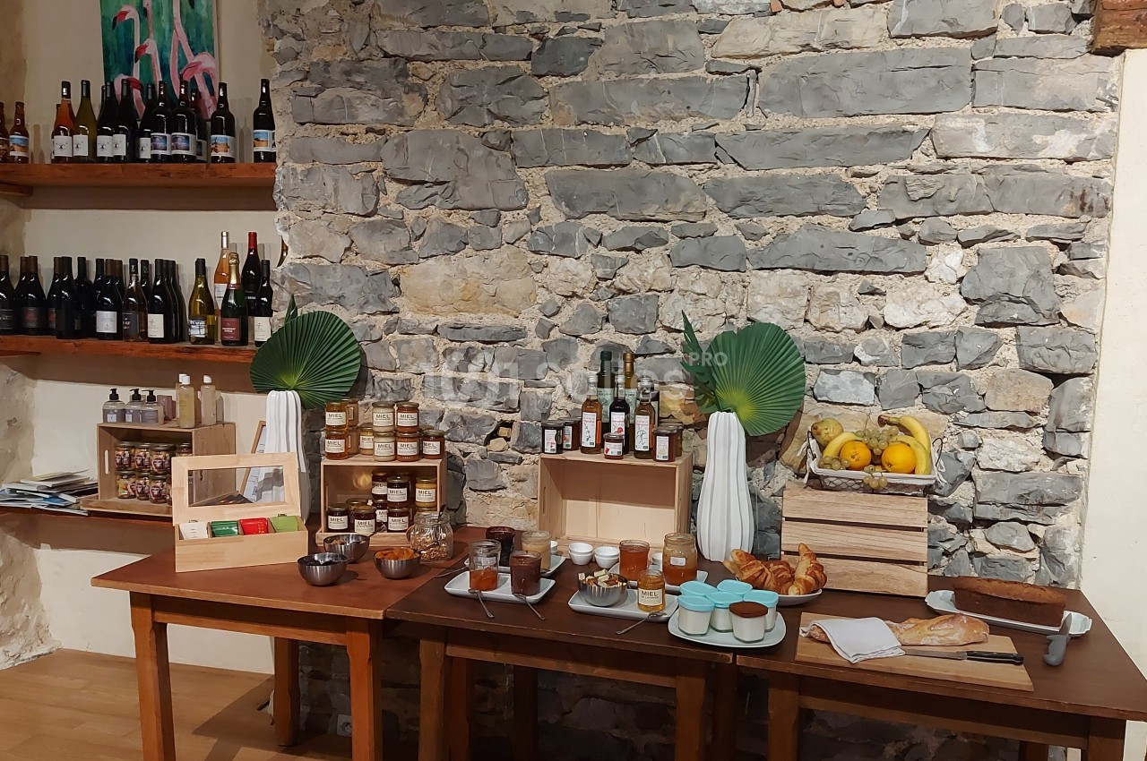 Table avec produits locaux : confitures, miel, pain, viennoiseries, fruits, boissons et décorations végétales devant un mur…