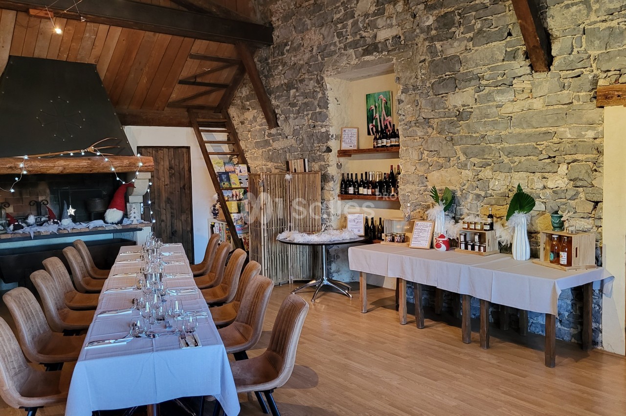 Salle rustique avec murs en pierre, grande table dressée, cheminée décorée et tables présentant des produits locaux.