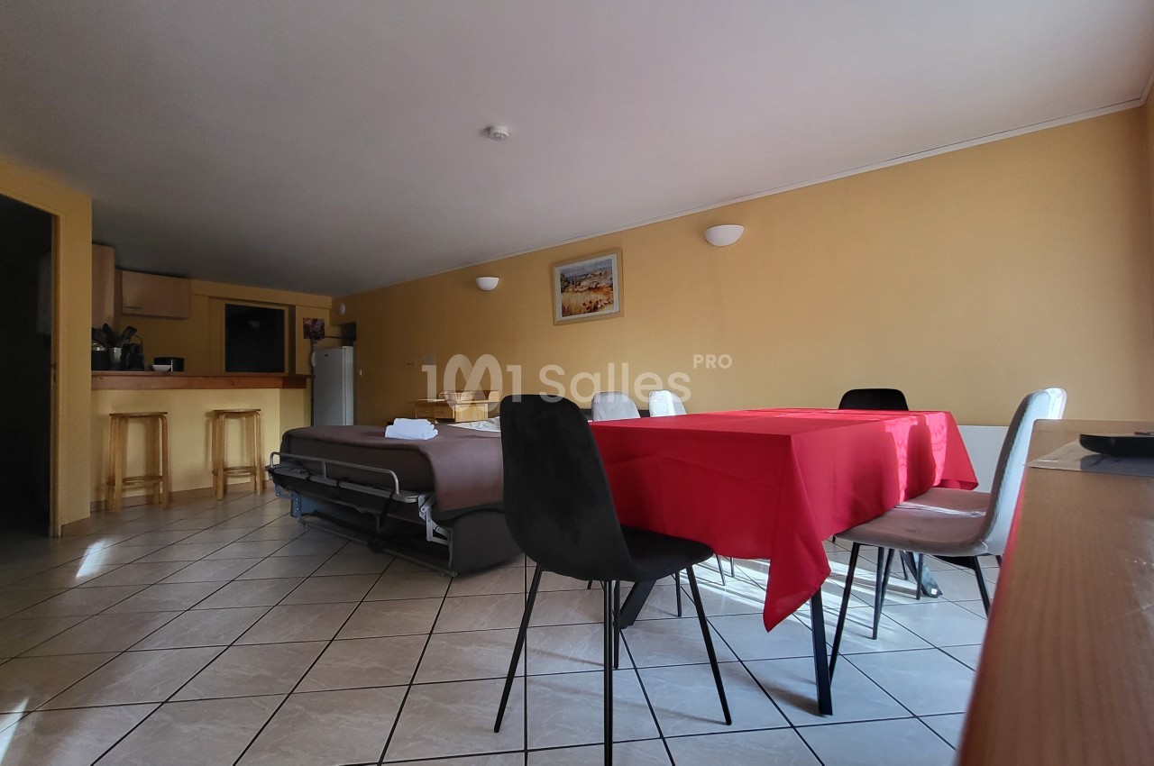 Salle à manger avec table recouverte d'une nappe rouge, chaises noires et blanches, cuisine ouverte et canapé-lit en arrière…