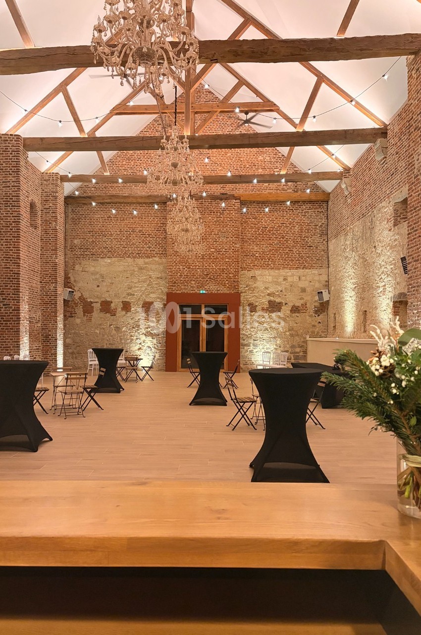 Salle en briques avec poutres apparentes, tables hautes noires et éclairage chaleureux, vue depuis un comptoir en bois.