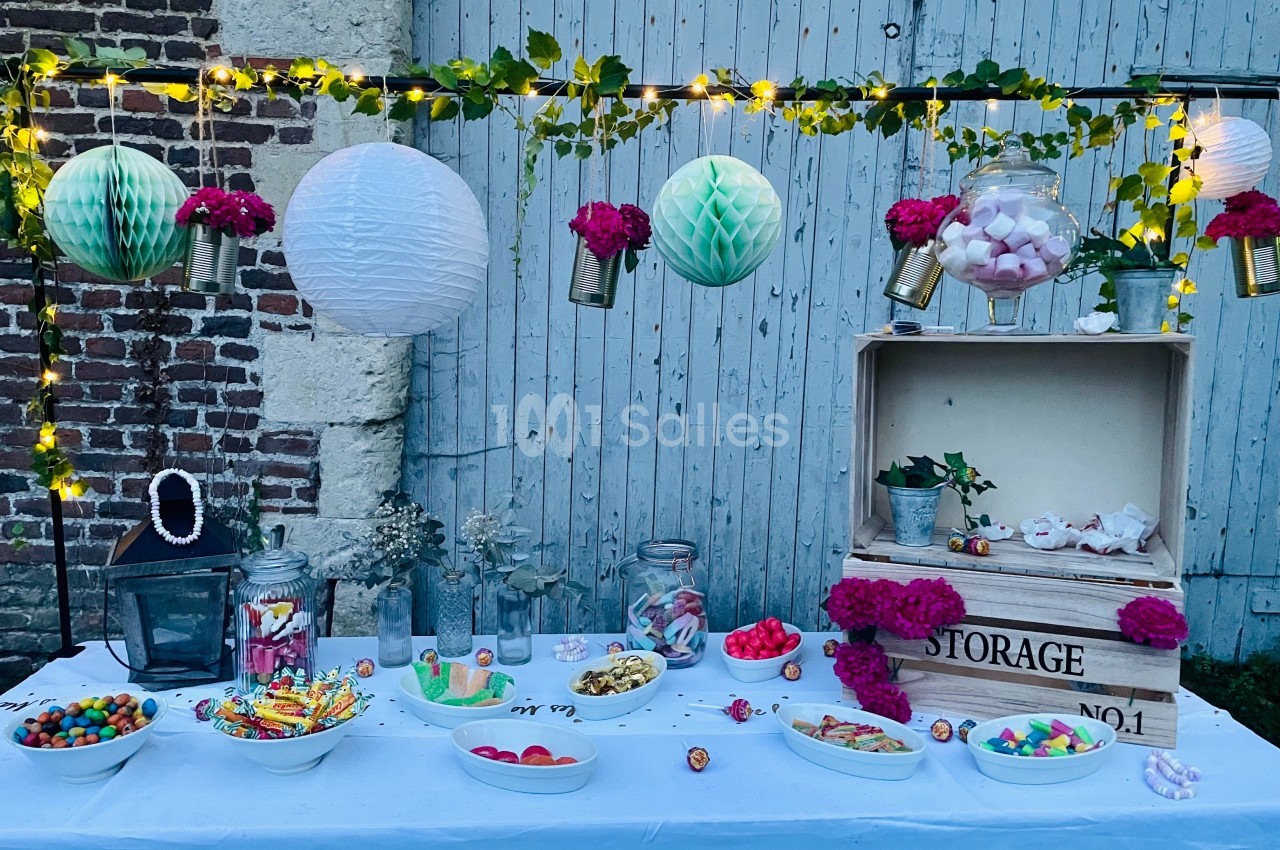 Table décorée pour une fête avec guirlandes lumineuses, fleurs, bonbons variés et lanternes suspendues devant un mur en bois.