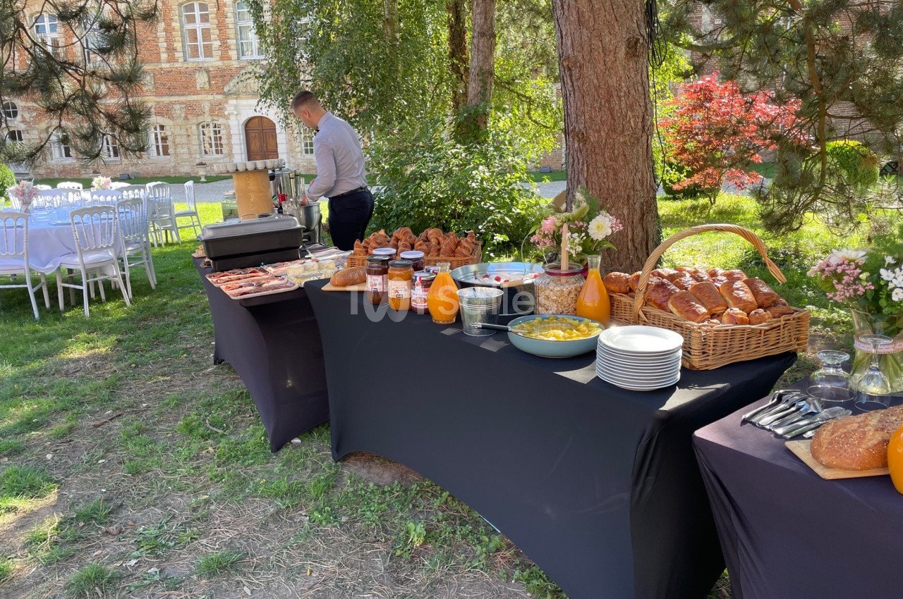 Buffet en plein air avec viennoiseries, jus, confitures et vaisselle, installé sous des arbres près d'un bâtiment en briques.