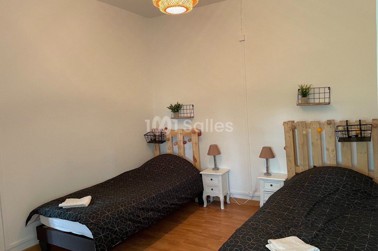 Deux lits simples avec linge de lit sombre, tables de chevet, lampes et décoration murale dans une chambre lumineuse.
