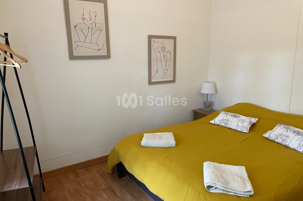 Chambre avec un lit double couvert d'une housse jaune, deux serviettes pliées et des dessins encadrés au mur.