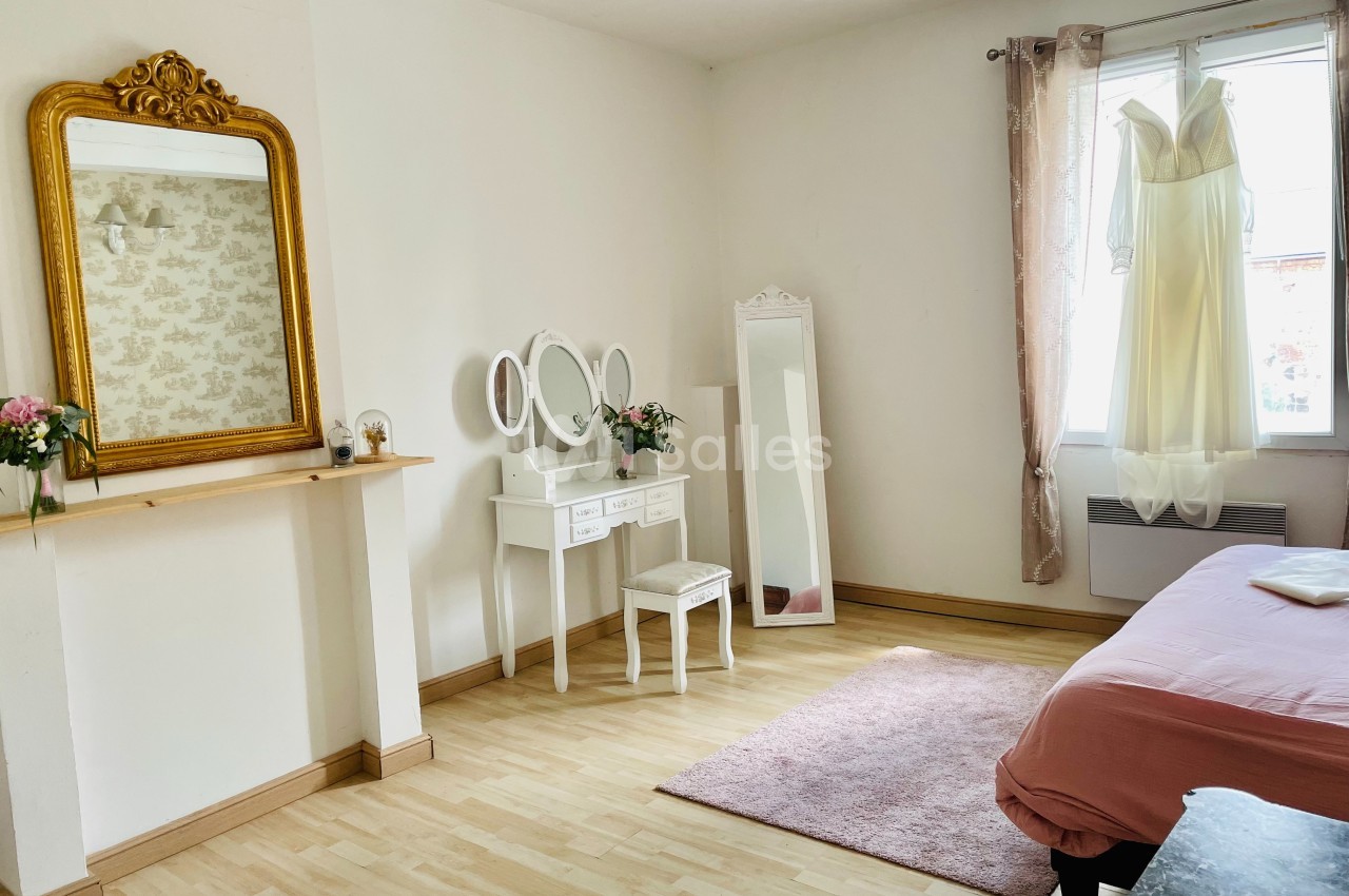 Chambre lumineuse avec coiffeuse blanche, miroir doré, robe suspendue à la fenêtre et lit avec couverture rose.