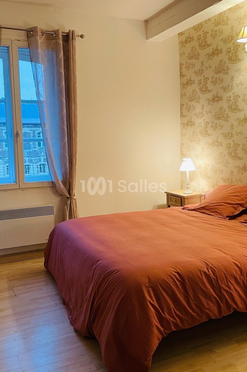 Chambre lumineuse avec un lit double aux draps orange, une lampe de chevet et une fenêtre donnant sur des bâtiments.