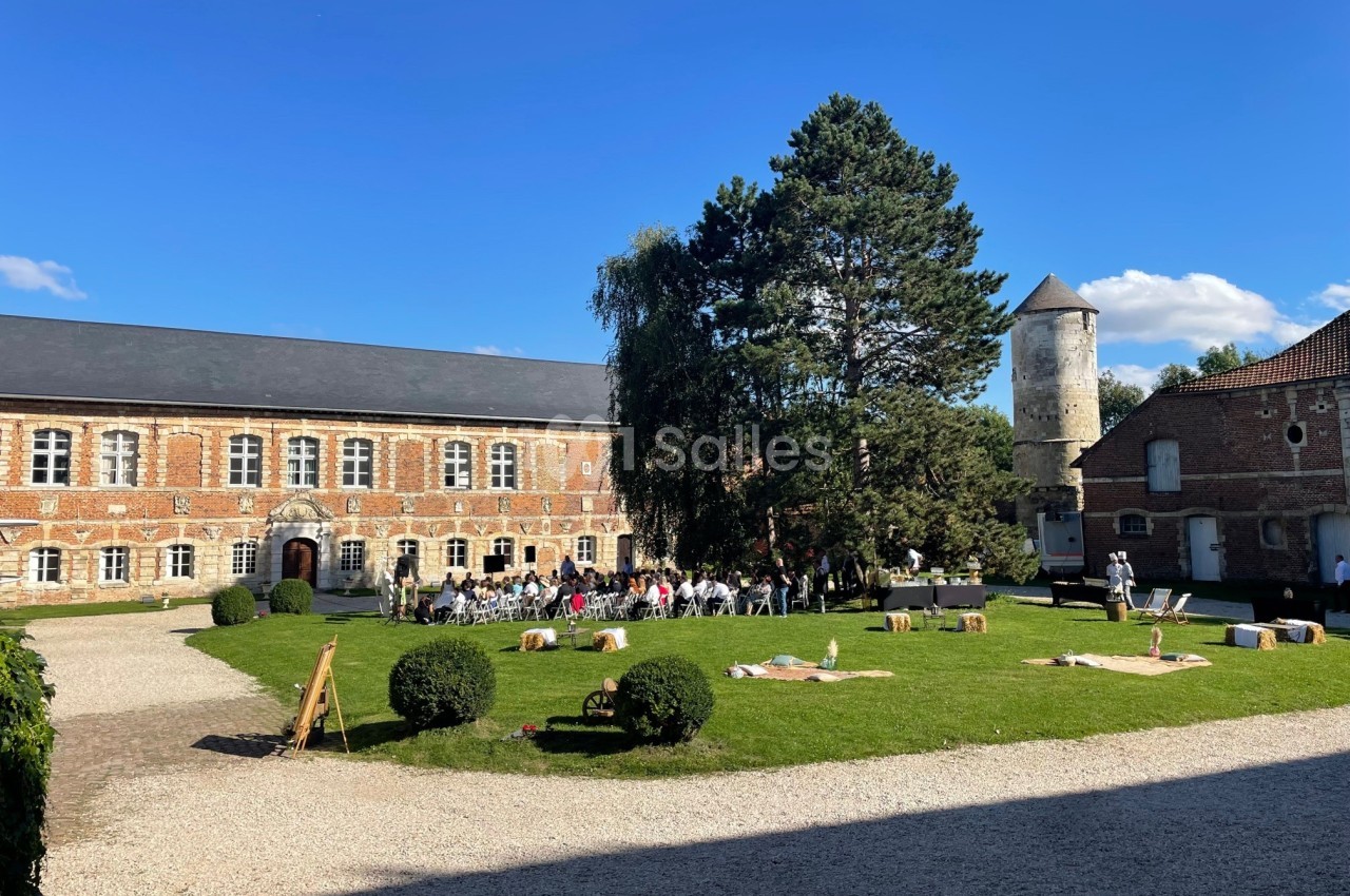 Cour d'une ancienne bâtisse en briques avec pelouse, arbres, tour ronde et invités rassemblés pour un événement.