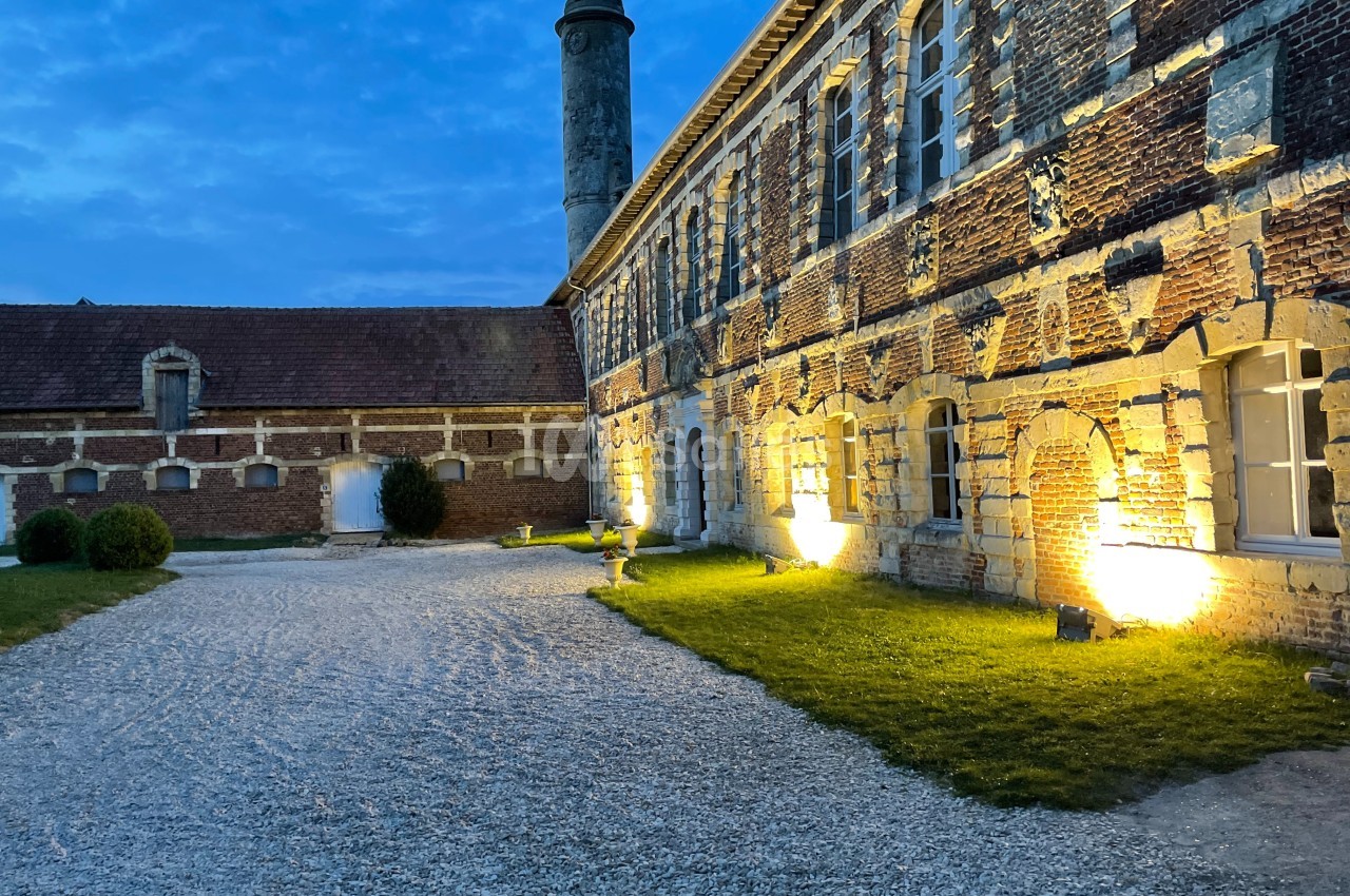 Cour pavée d'un bâtiment en briques anciennes, éclairé par des lumières au crépuscule, avec une cheminée en arrière-plan.