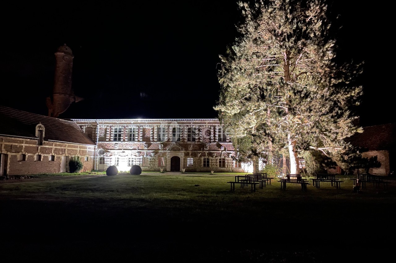 Façade d'un bâtiment ancien éclairé la nuit, entouré d'arbres et de tables sur une pelouse.