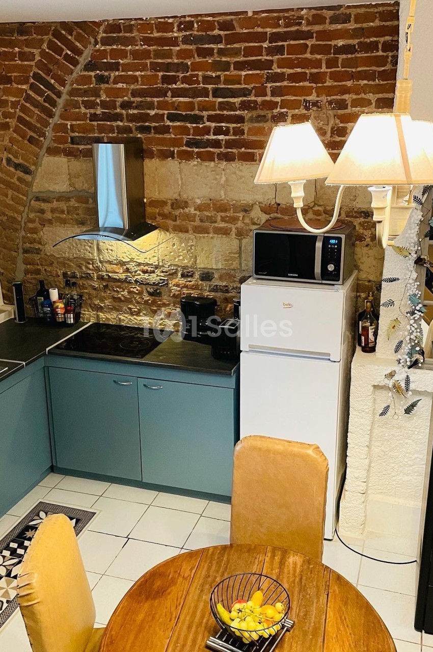 Cuisine avec murs en briques apparentes, meubles bleu-gris, table ronde en bois, réfrigérateur blanc et éclairage suspendu.